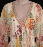 A cream-colored floral long-sleeve top with a V-neckline, featuring yellow, orange, and red flowers with green leaves and a gold embroidered trim, displayed on a mannequin.