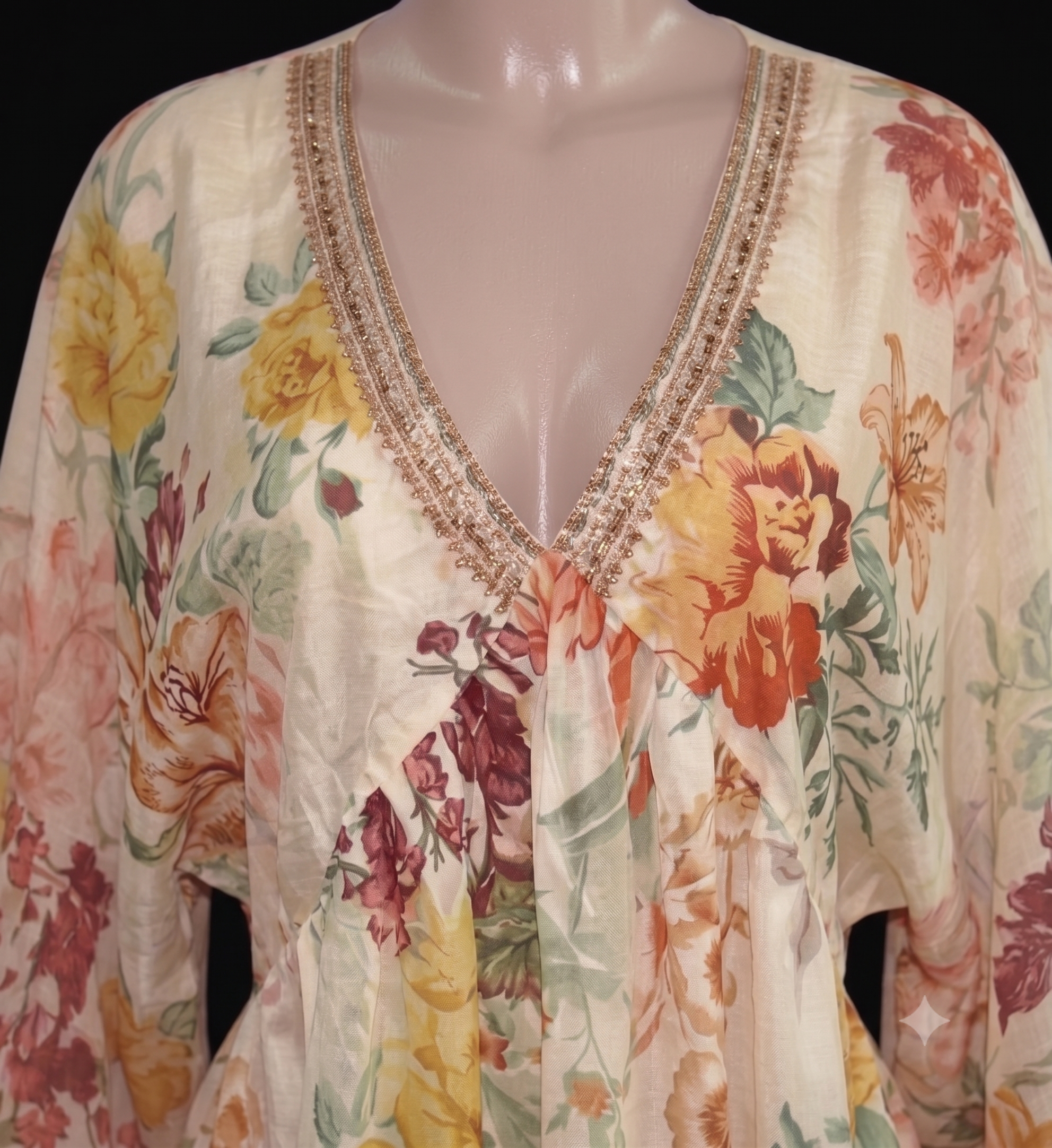 A cream-colored floral long-sleeve top with a V-neckline, featuring yellow, orange, and red flowers with green leaves and a gold embroidered trim, displayed on a mannequin.