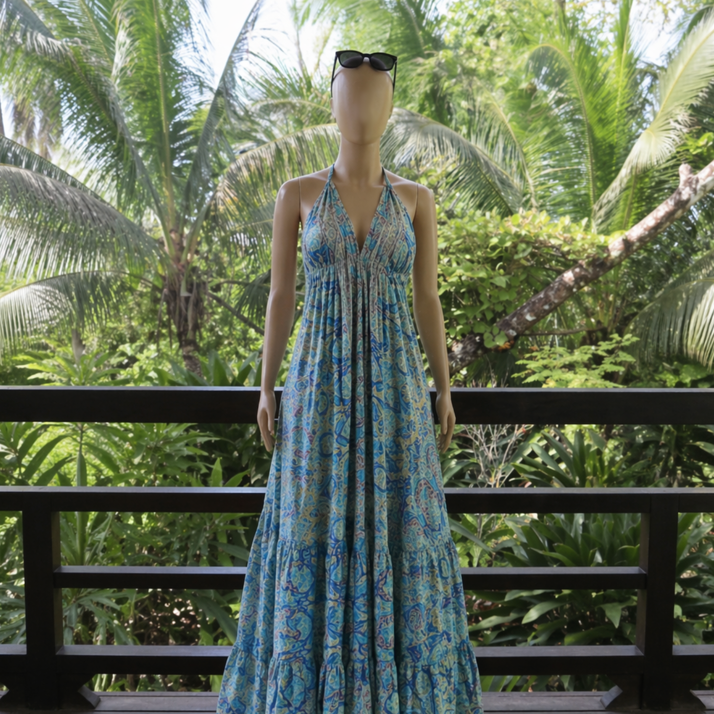 Mannequin wearing a blue floral dress with sunglasses on a wooden deck surrounded by tropical plants.