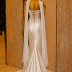 White wedding dress with lace back and cape on a mannequin against a beige wall.