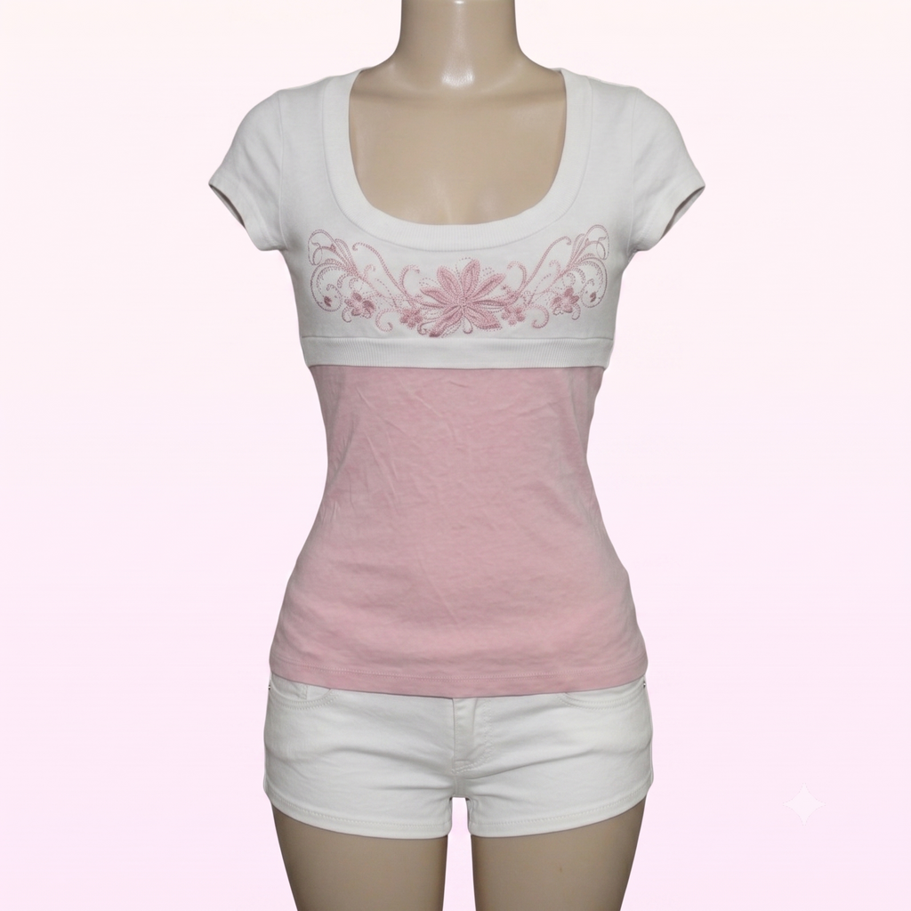A women's short-sleeve scoop-neck top displayed on a mannequin, featuring a white upper section with pink floral embroidery across the chest and a solid light pink lower section, paired with white shorts.