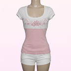 A women's short-sleeve scoop-neck top displayed on a mannequin, featuring a white upper section with pink floral embroidery across the chest and a solid light pink lower section, paired with white shorts.