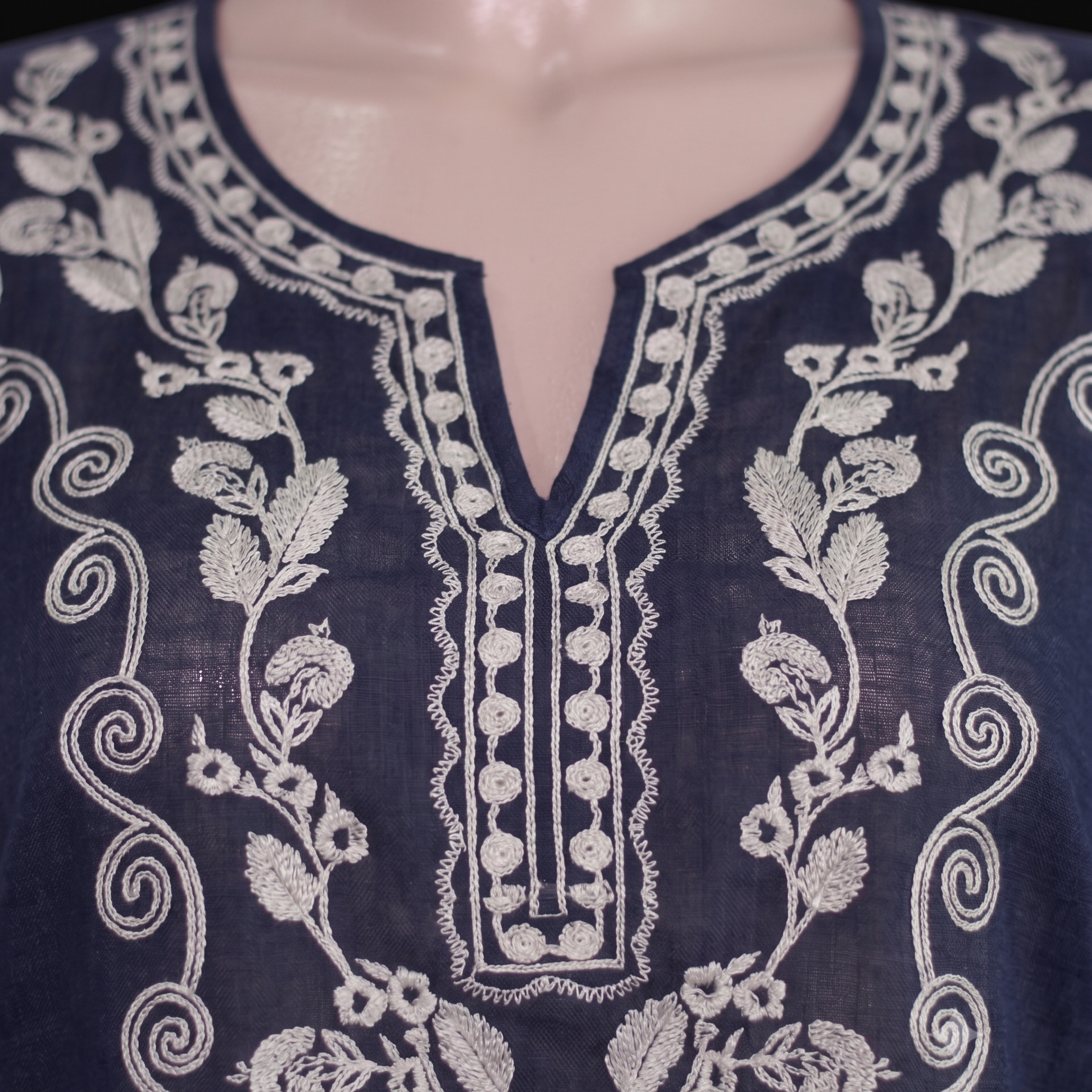 Close-up of a navy blue embroidered tunic top displayed on a mannequin torso, featuring intricate white floral and scroll embroidery around a split neckline.